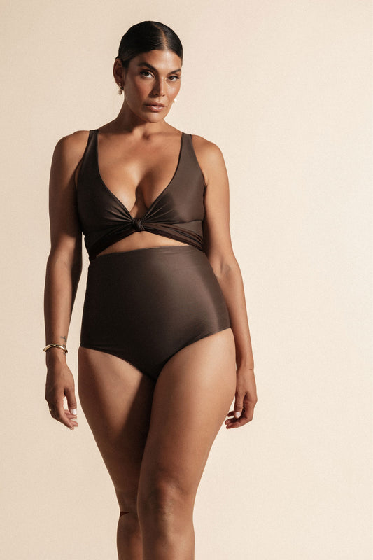 Woman stands in front of a beige background wearing a deep chocolate brown coloured bikini set. The top is cinched at the bust with a tie and the bottoms are high cut.