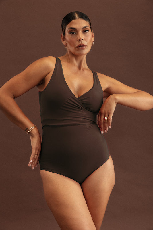 A woman stands in front of a soft brown backdrop whilst wearing a sculpting wrap swimsuit in a rich, deep chocolate colour.