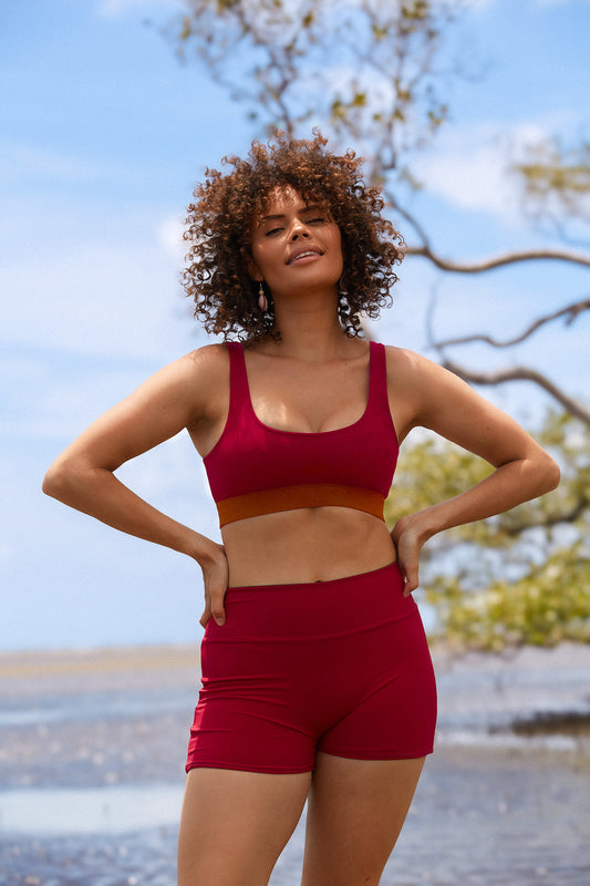 Woman stands on a beach wearing a raspberry coloured swim set featuring high waisted shorts and a sports bra shaped swim top.