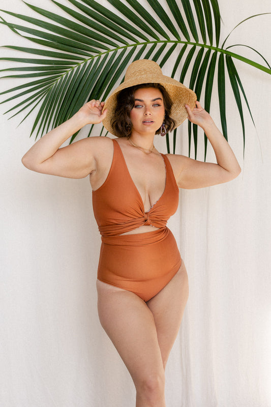 Woman wearing a terracotta orange bikini set stands adjusting her straw hat in front of a palm frond.
