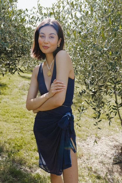 Woman posing in garden wearing a dark blue bikini top, with blue sarong tied around her waist.