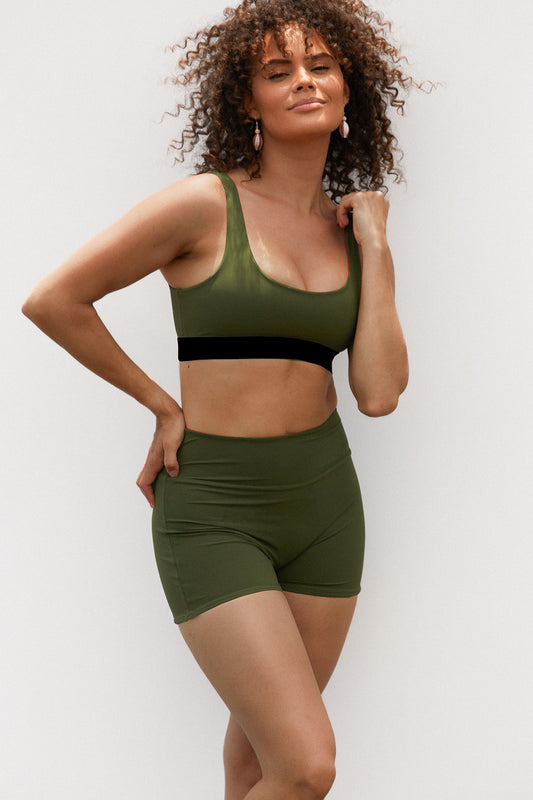 Woman wears a eucalypt coloured swim set with high waisted shorts and a sports bra shaped swim top. The top has thick straps and a black coloured band underneath the bust.