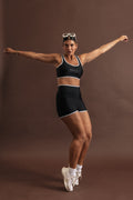 Woman wearing a black swim set strikes an action pose, standing on the ends of her toes. The bottoms are high waisted and top is a scoop neck, sports bra shape. Both have white binding-style trim on the outer edges.