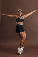 Woman wearing a black swim set strikes an action pose, standing on the ends of her toes. The bottoms are high waisted and top is a scoop neck, sports bra shape. Both have white binding-style trim on the outer edges.