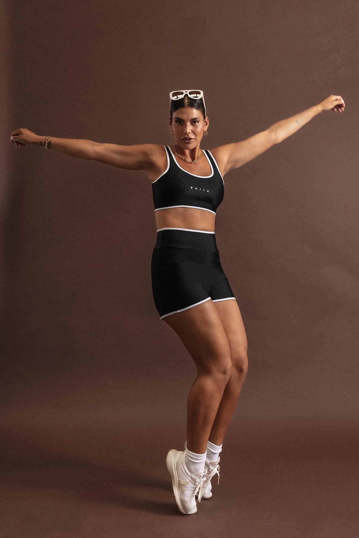 Woman wearing a black swim set strikes an action pose, standing on the ends of her toes. The bottoms are high waisted and top is a scoop neck, sports bra shape. Both have white binding-style trim on the outer edges.
