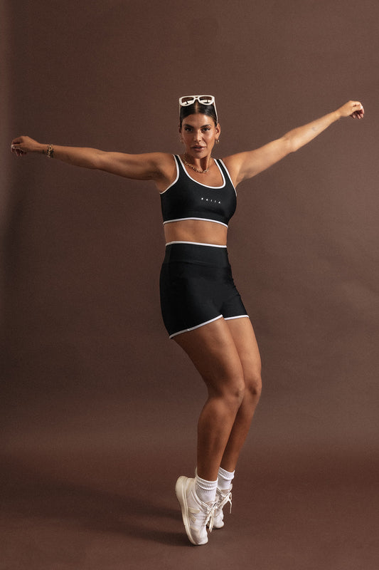 Woman wearing a black swim set strikes an action pose, standing on the ends of her toes. The bottoms are high waisted and top is a scoop neck, sports bra shape. Both have white binding-style trim on the outer edges.