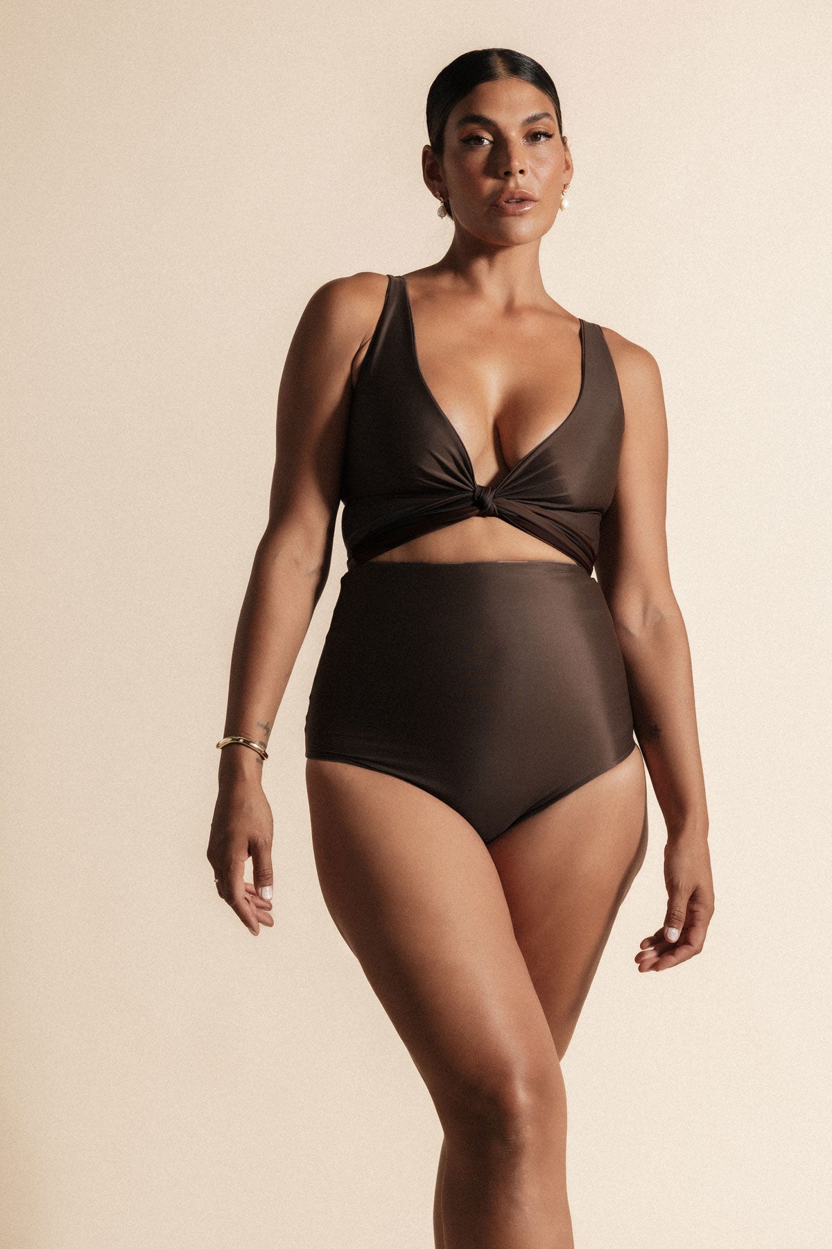 Woman stands in front of a beige background wearing a deep chocolate brown coloured bikini set. The top is cinched at the bust with a tie and the bottoms are high cut.