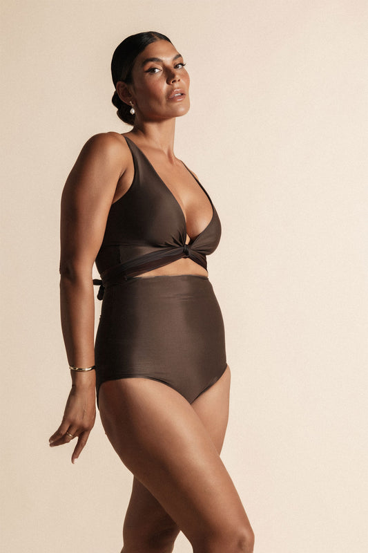 Woman stands in front of a beige background wearing a deep chocolate brown coloured bikini set. The top is cinched at the bust with a tie and the bottoms are high cut.