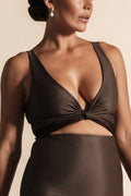 Woman stands in front of a beige background wearing a deep chocolate brown coloured bikini set. The top is cinched at the bust with a tie and the bottoms are high cut.