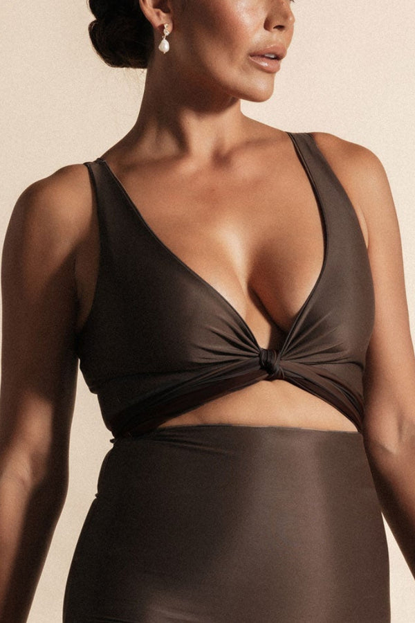 Woman stands in front of a beige background wearing a deep chocolate brown coloured bikini set. The top is cinched at the bust with a tie and the bottoms are high cut.
