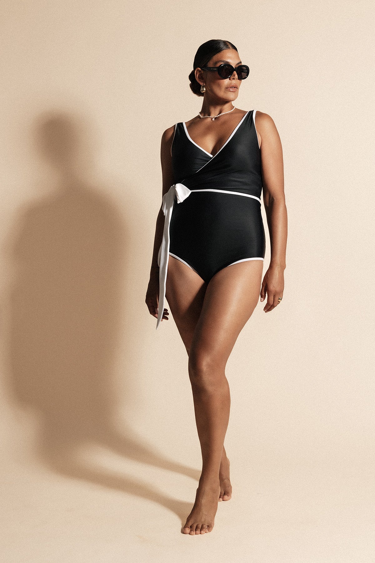 Woman stands with lighting silhouetting her shadow onto the beige backdrop behind. She wears a solid black coloured Woman stands with lighting silhouetting her shadow onto the beige backdrop behind. She wears a solid black coloured contouring wrapsuit framed by a white waist tie and binding. The look is completed with luxurious sunglasses as she gazes toward the light source.