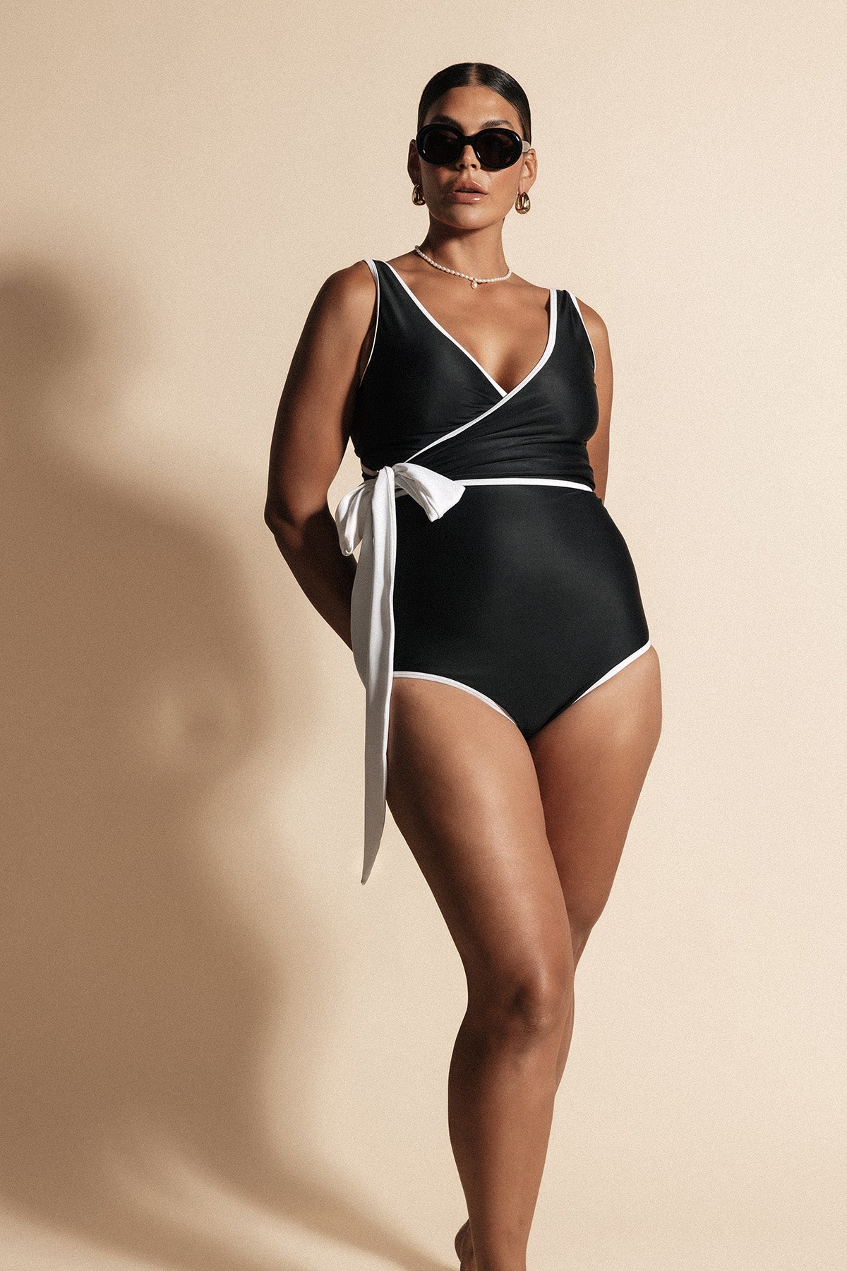 Woman stands with lighting silhouetting her shadow onto the beige backdrop behind. She wears a solid black coloured contouring wrapsuit framed by a white waist tie and binding. The look is completed with luxurious black sunglasses as she gazes toward the light source.