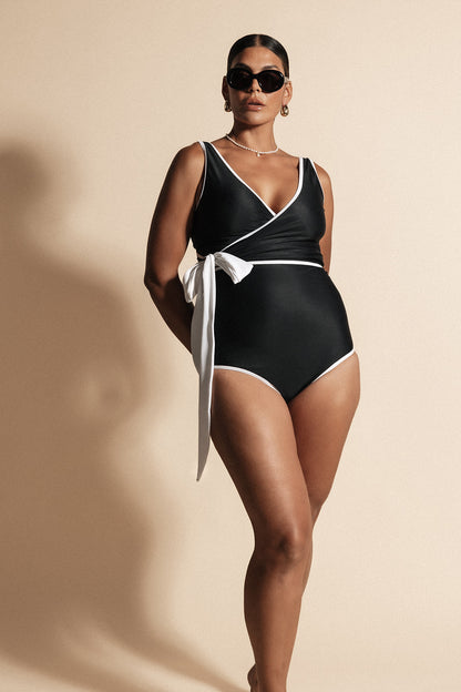 Woman stands with lighting silhouetting her shadow onto the beige backdrop behind. She wears a solid black coloured contouring wrapsuit framed by a white waist tie and binding. The look is completed with luxurious black sunglasses as she gazes toward the light source.
