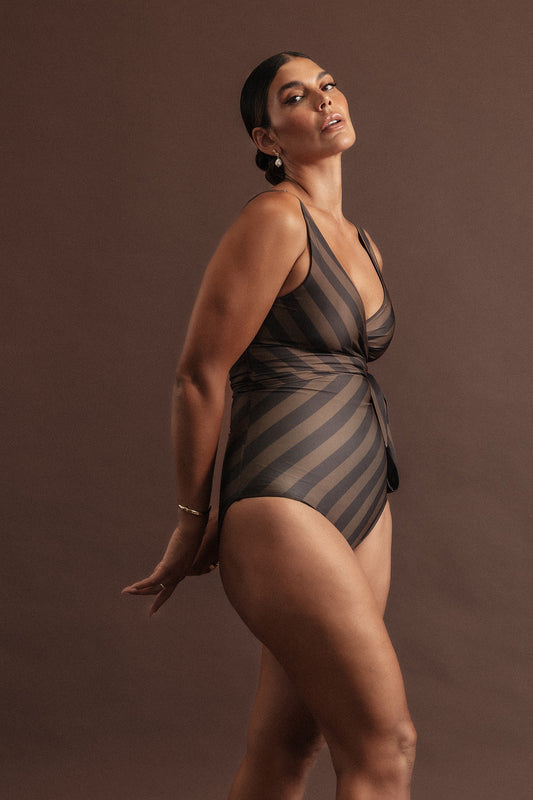 A woman posing in front in of an earthy brown backdrop with her hands behind her back, wearing a sculpting wrapsuit with deep chocolate and light macchiato brown stripes.