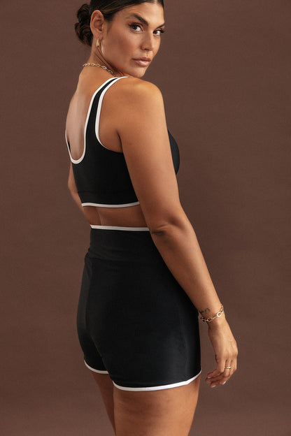Shot of a model from behind wearing a swim set, showcasing the back coverage of the Andi swim top and the sculpting properties of the high waisted Andi shorts. The set is black with white binding trim.