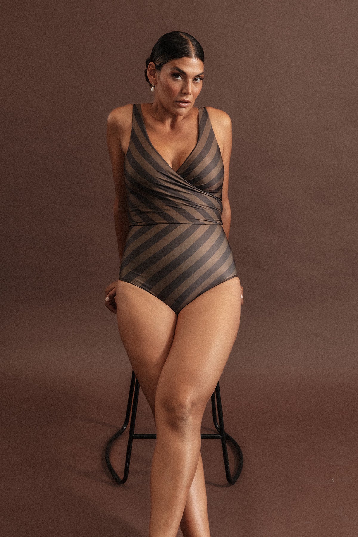 Woman leans on a stool wearing a deep chocolate and macchiato coloured bikini set with a bold diagonal stripe pattern.