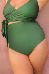 Woman leaning against wall, wearing a juniper green 3 piece bikini.