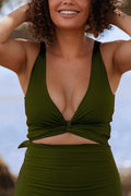 A woman standing on a beach wearing a deep green bikini set smiles as she runs her hands through her hair. 