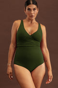 Woman adorned with gold jewellery wearing a solid deep green wrapsuit in front of a brown studio background.