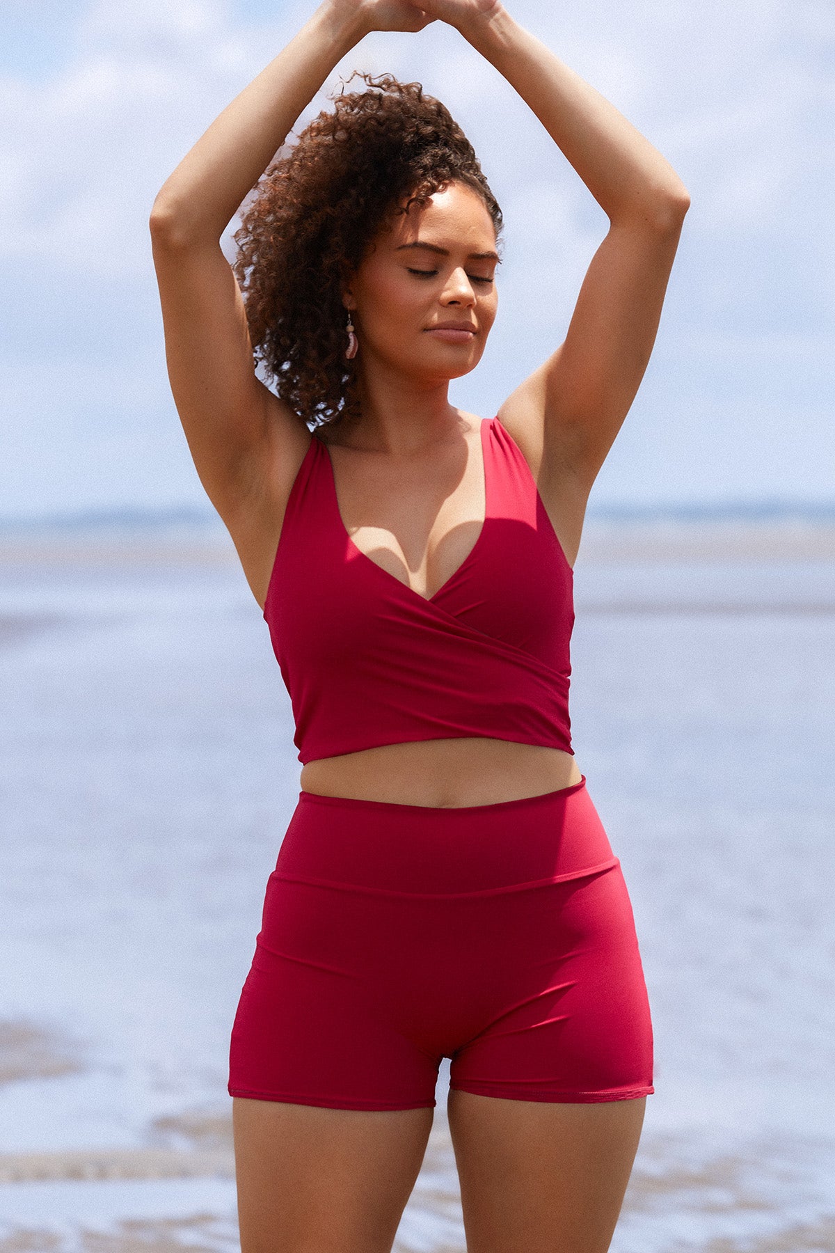 Woman stands on a beach wearing a raspberry coloured swim set featuring high waisted shorts and a two piece wrap bikini top.