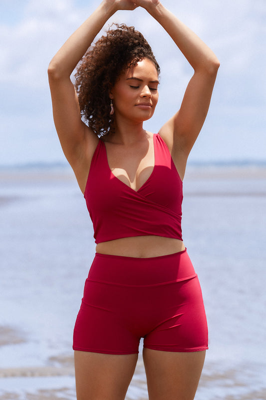 Woman stands on a beach wearing a raspberry coloured swim set featuring high waisted shorts and a two piece wrap bikini top.