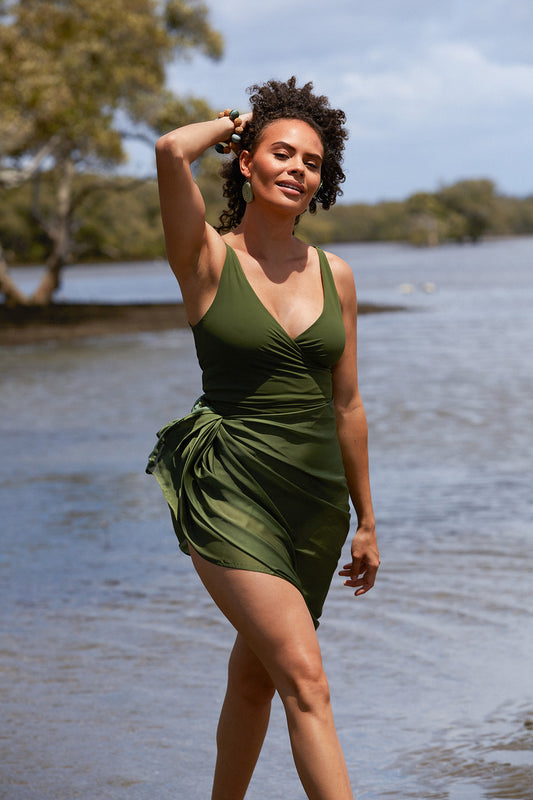 A woman brushing her hand through her hair in front of a lake, wearing a solid deep green wrap swimsuit. Her lower half is covered down to the knees by a matching green sarong tied around her waist.