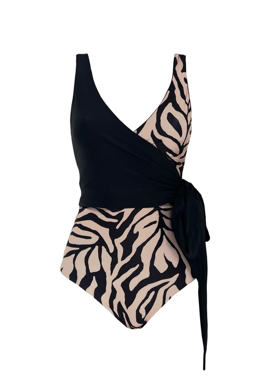 A woman stands with one hand on her hip, wearing a wrapsuit with a unique take on animal print featuring hints of cream palm detailing on a black base.