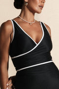 A woman stands against a beige backdrop wearing an all black bikini set with white binding.
