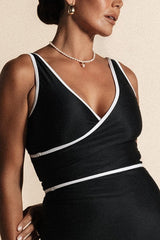 A woman stands against a beige backdrop wearing an all black bikini set with white binding.
