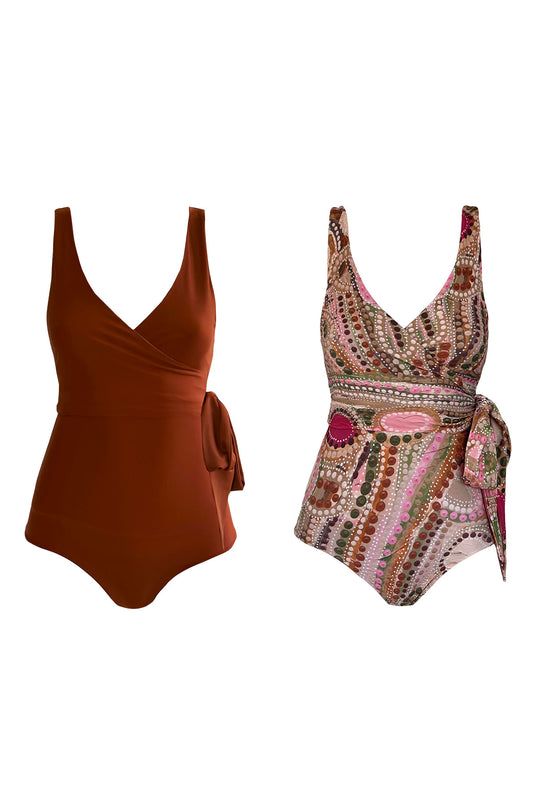 Woman wears wrapsuit with indigenous artwork print featuring shades of rust and khaki, complemented by vibrant hues of raspberry pink. The swimsuit is complemented by a matching rust coloured waist tie, worn on the right hip in a bow.