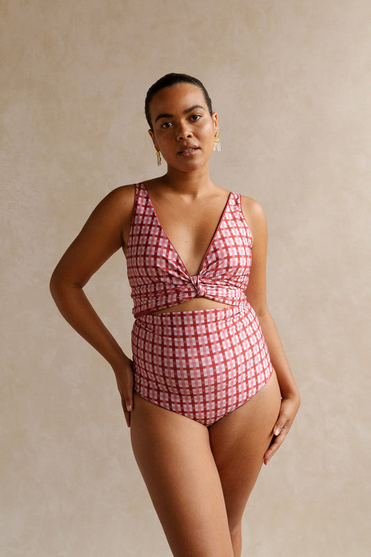Woman wearing a deep, burnt coral pink coloured gingham print bikini set with her hand on her hip standing in front of a beige studio backdrop. 