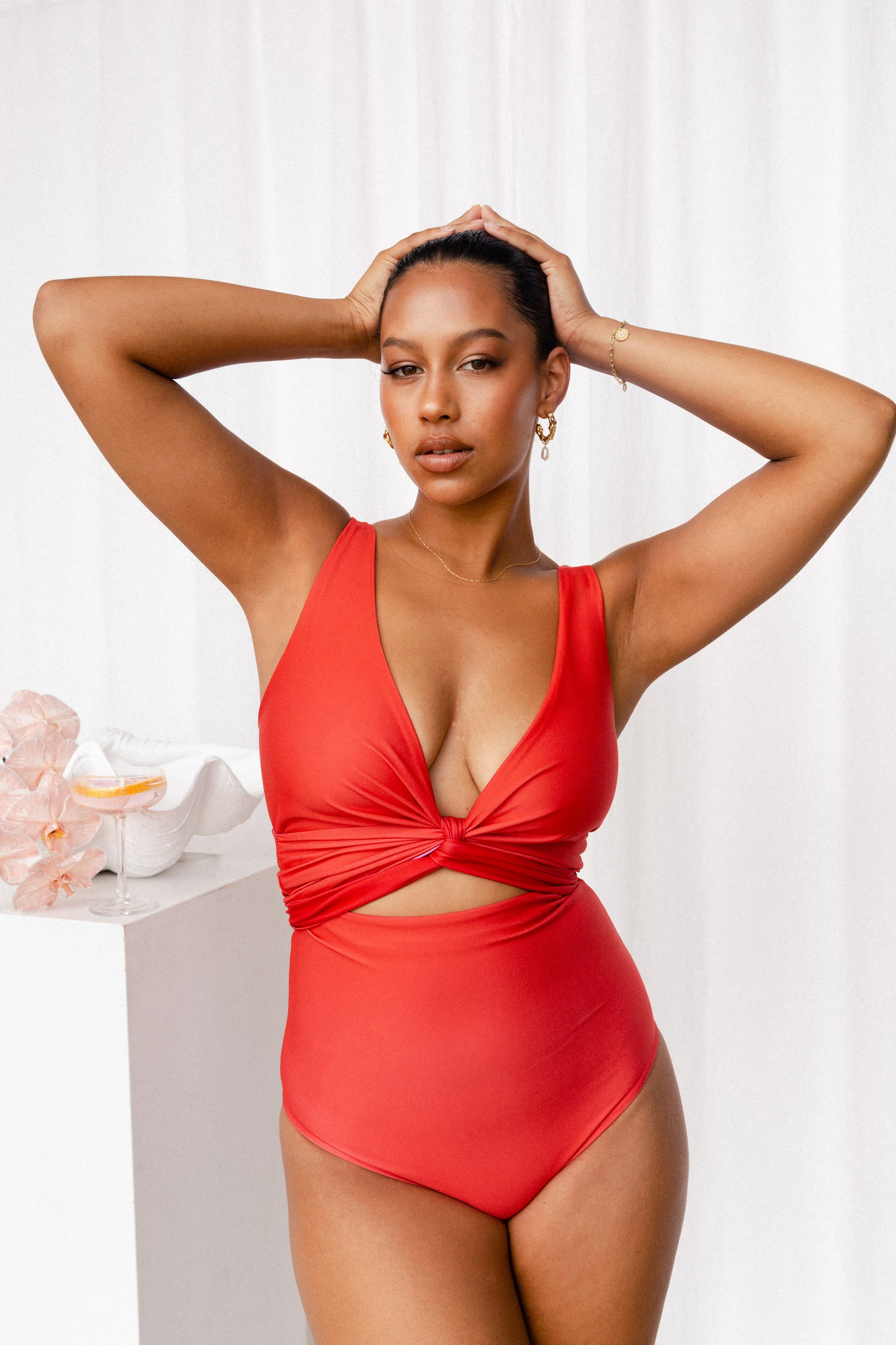 Deep coral red bikini set on a model who stands next to a white plinth adorned with shells and a cocktail.