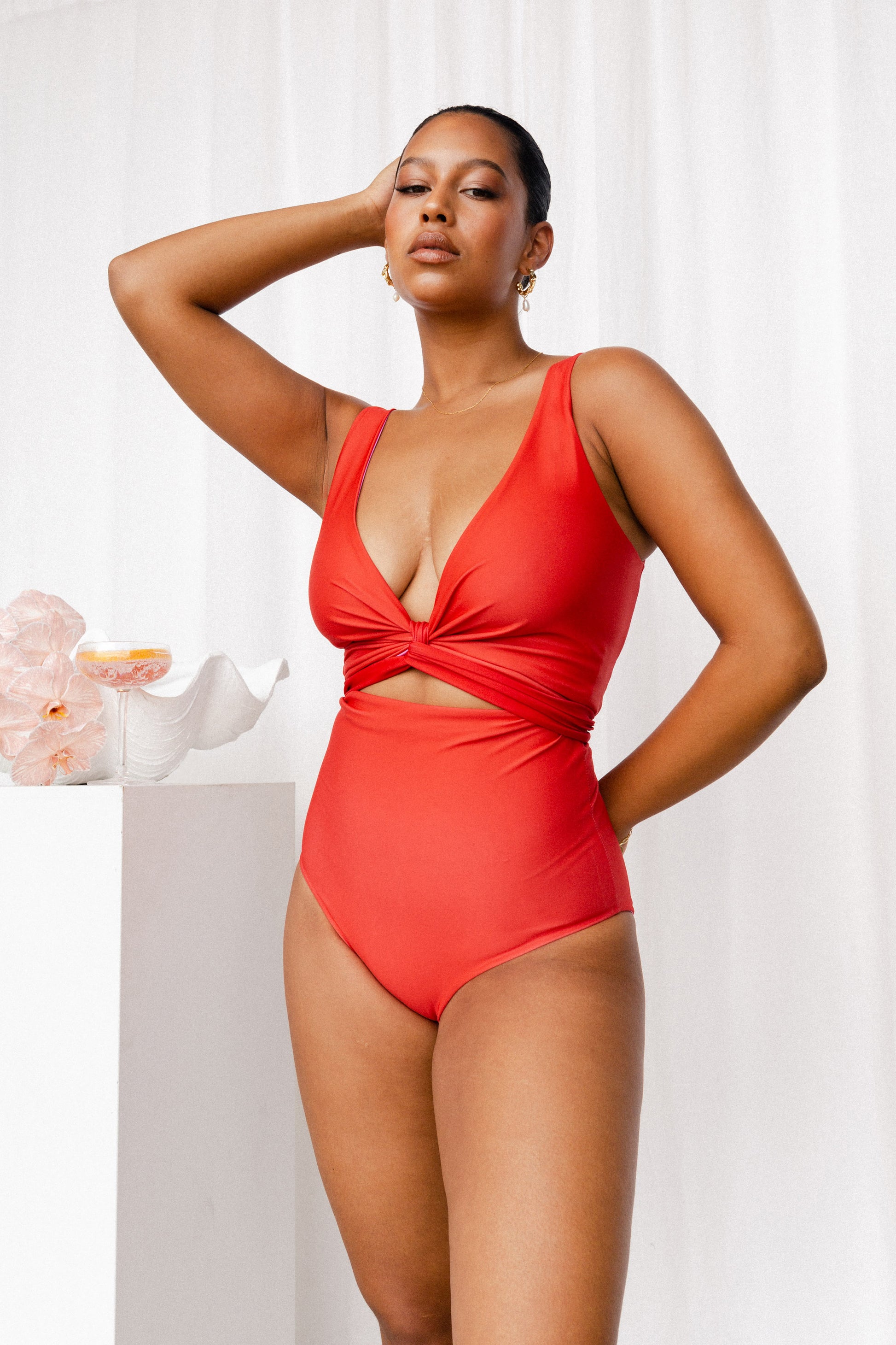 Deep coral red bikini set on a model who stands next to a white plinth adorned with shells and a cocktail.