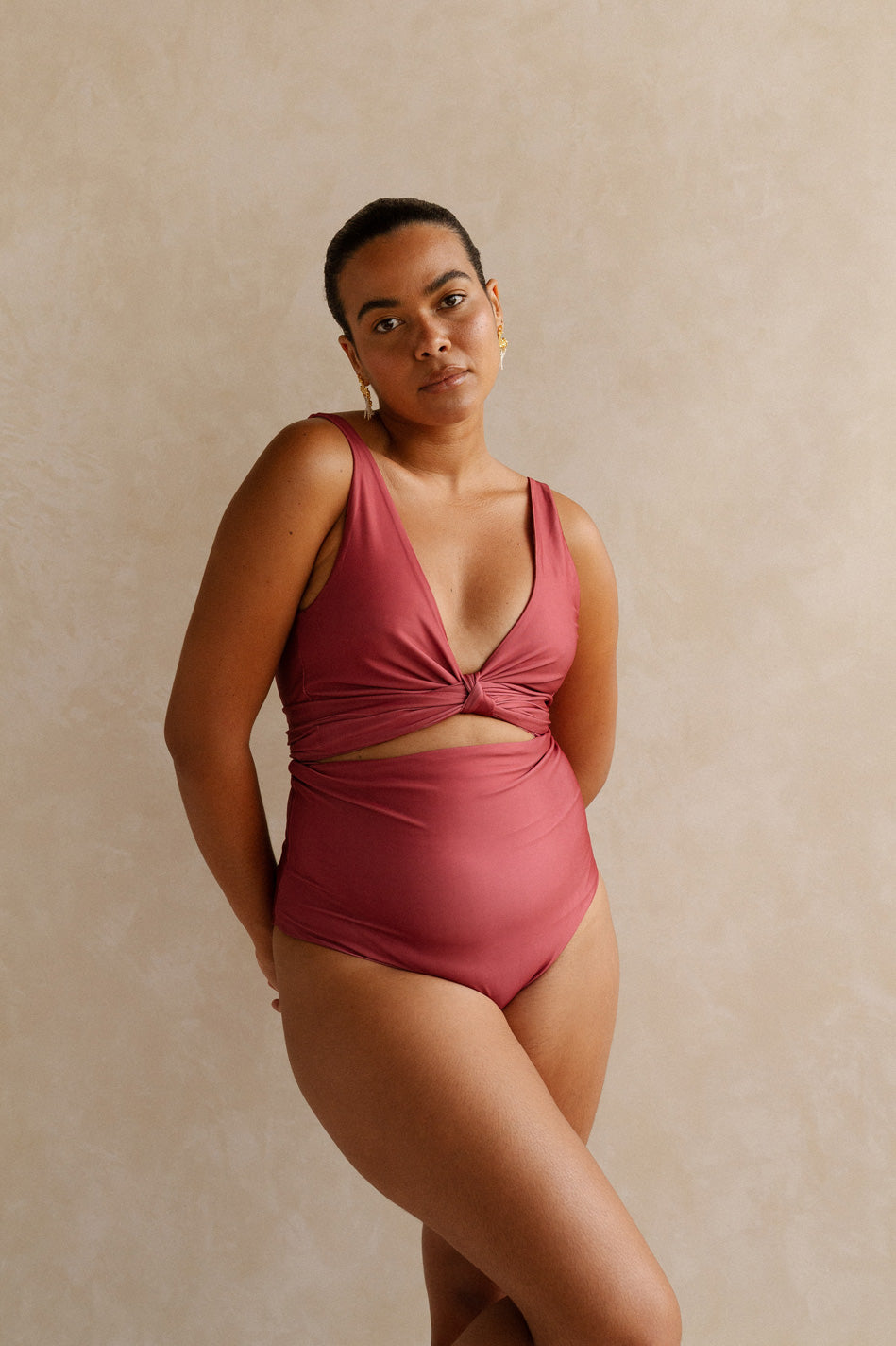 A woman standing with hands behind her back wears a deep, burnt coral pink bikini set. It has high waisted bottoms and is cinched at the bust to provide a plunging neckline.