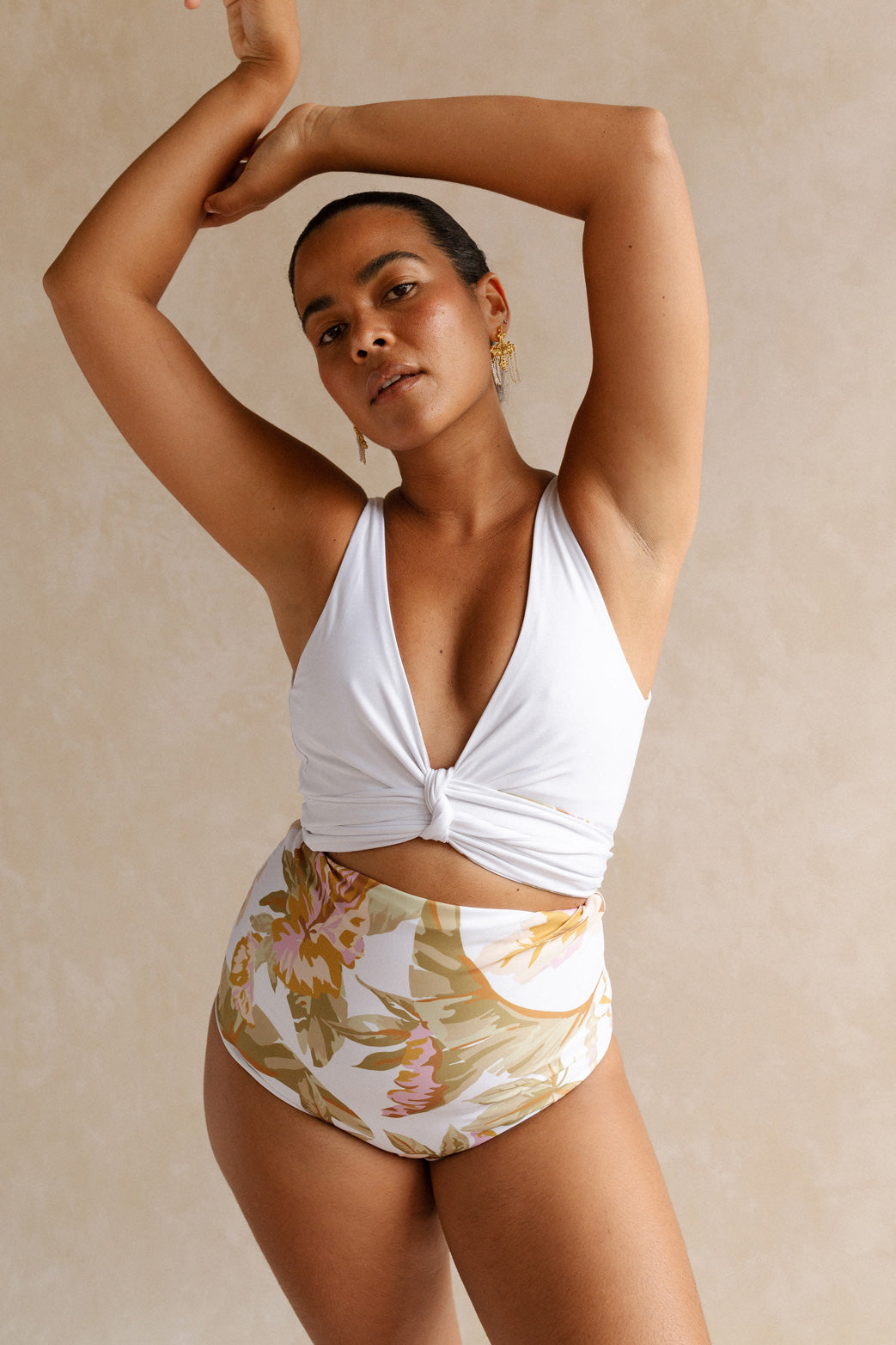 Woman stands in front of beige background with arms raised above her head. She wears a solid white bikini top cinched at the bust to form a flattering low cut. It is paired with matching high waisted bikini bottoms, reversed to showcase the gold, sage and pink tones of the floral print side.