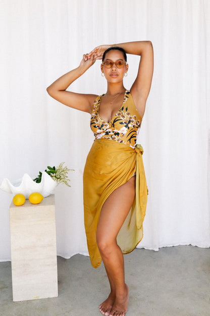 Woman poses in a mustard printed bikini set with matching sarong tied at the waist.