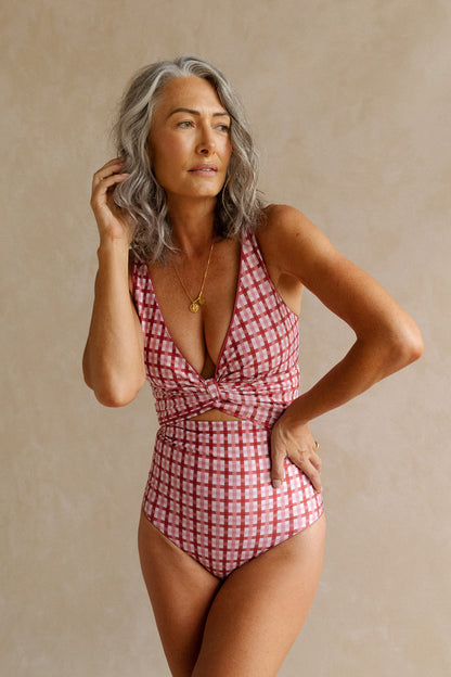 Woman wearing a deep, burnt coral pink coloured gingham print bikini set stands in front of a beige studio backdrop. 