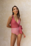 Woman stands in front of a matte beige backdrop wearing a wrapsuit featuring a coral pink gingham pattern. One chest piece has been reversed to showcase the solid pink colour, with a matching bow tied at the hip.