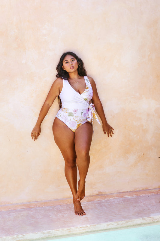 Woman laying against a stone wall by the pool dressed in the colourful vintage post-card motifs printed Havana reversible wrapsuit 