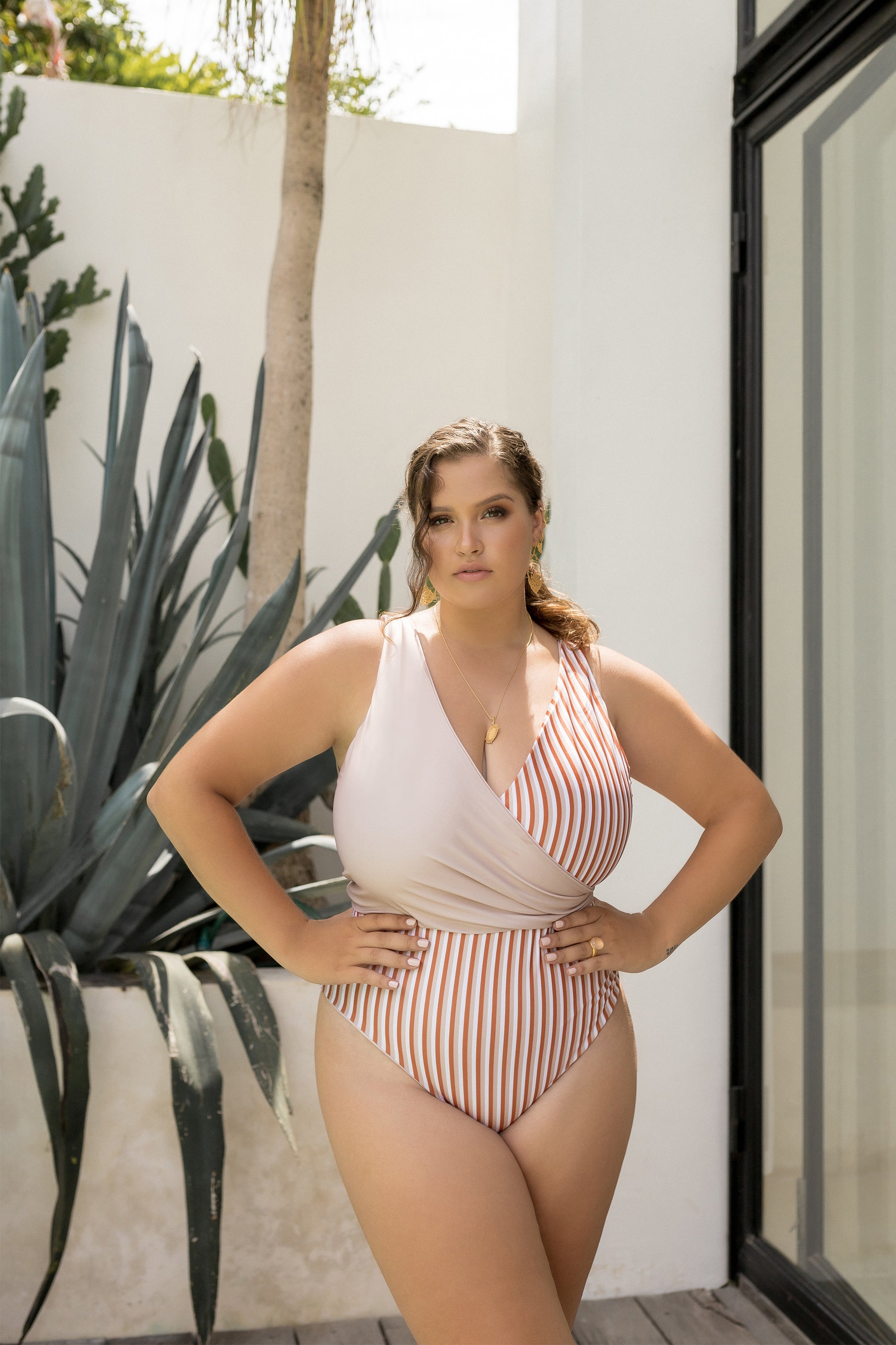 Woman stands in front of agave plant wearing a striped wrap swimsuit featuring rust, white & champagne-hued awning stripes. One chest piece is reversed to show the solid champagne side.