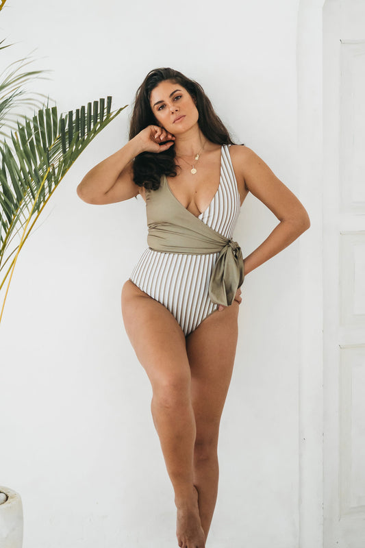 A woman stands beside palm frond wearing a wrap swimsuit with a creamy palette of clean white, stone and olive stripes. The chest piece is reversed to showcase the soft khaki side matching the stripes of the patterned side.
