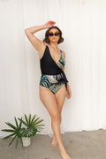 Woman stands with hand on her head wearing a hand-painted floral print swimsuit. The swimsuit features bold pink and green tones on a black base.