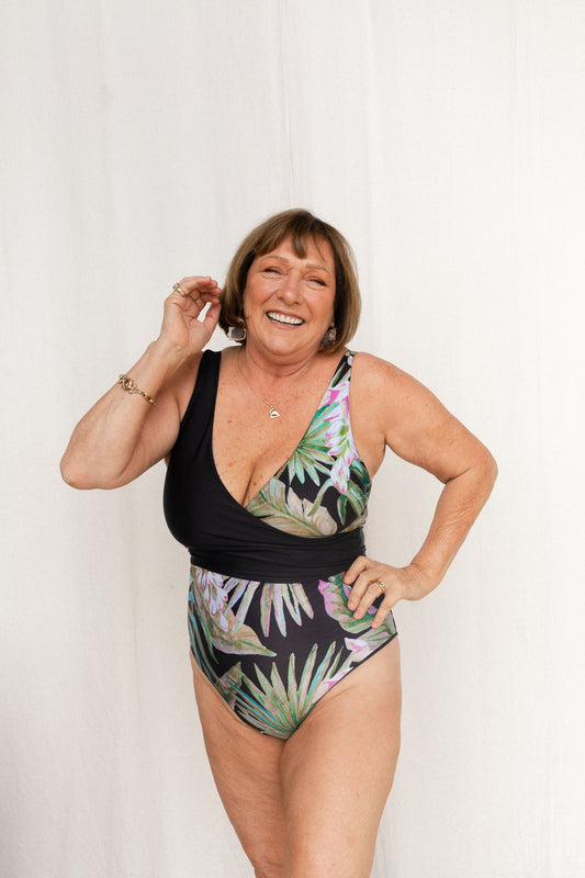 Woman stands in front of white curtain wearing a hand-painted floral print wrapsuit. The bold green and pink tones of the print are emphasised on a black base.