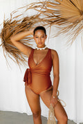 A woman posing in front of a white curtain and dried palm leaves, wearing a warm bronze coloured wrapsuit. 